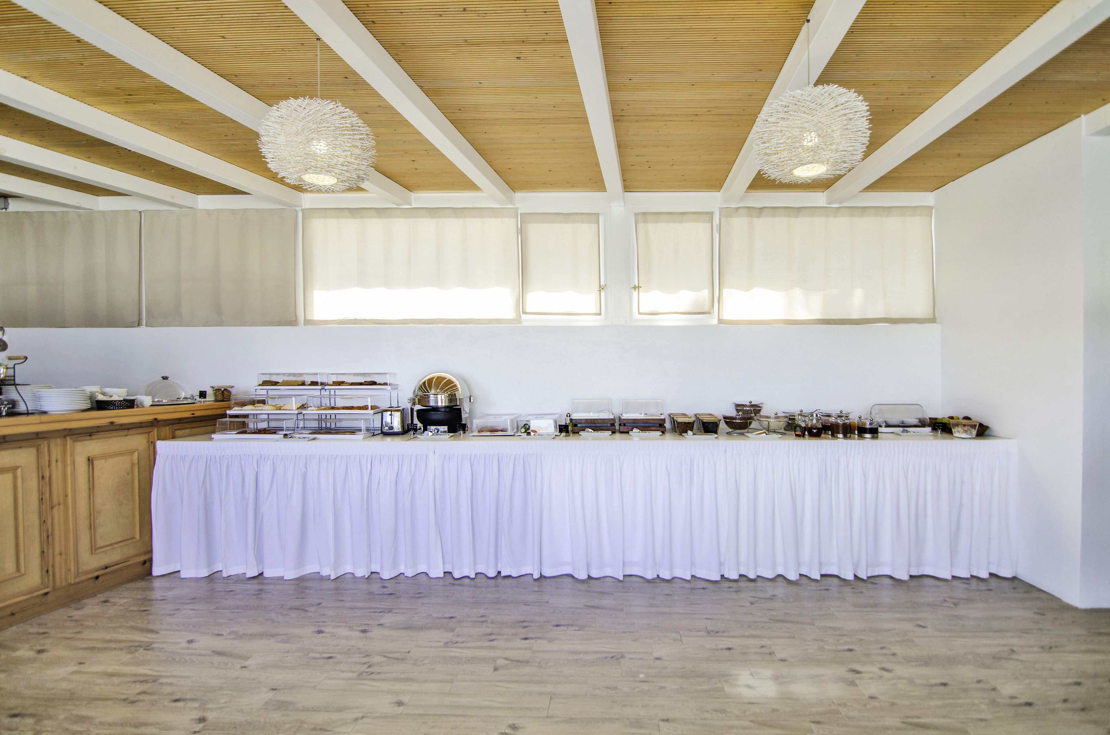 A spacious room with a long table covered in white cloth, wooden beams on the ceiling, and large windows with curtains.