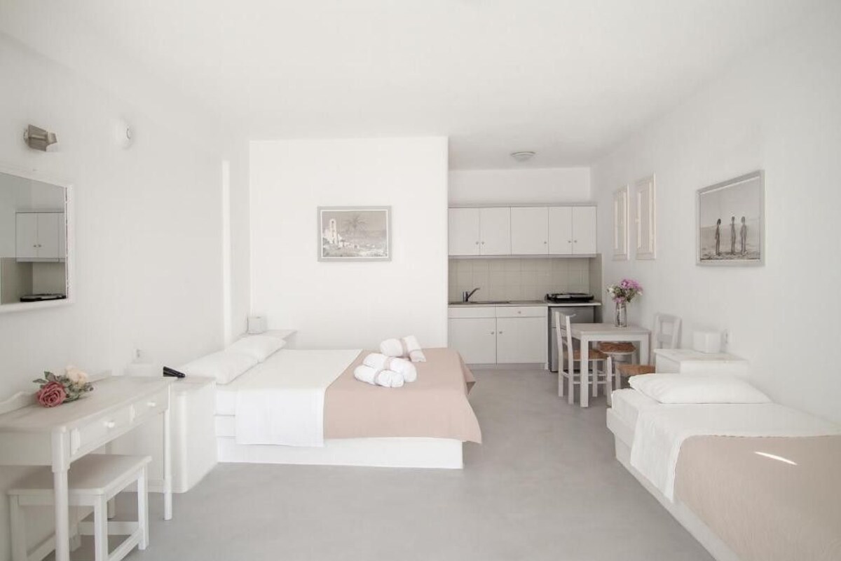 A minimalist hotel room with a bed, a desk, a small table, and a kitchenette.