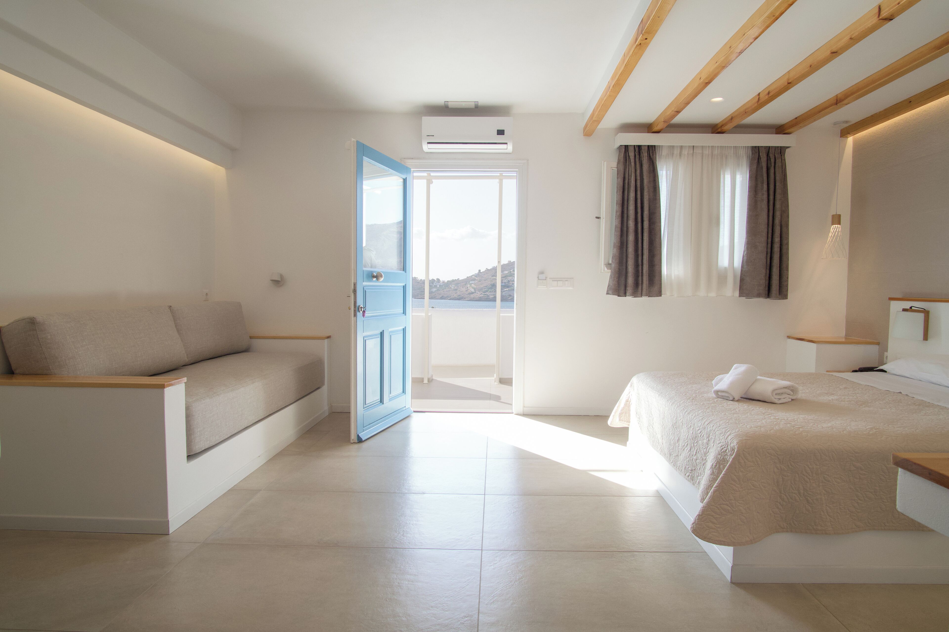Een moderne hotelkamer met een bank, een bed met een beige deken, een balkon met uitzicht en een houten plafond.