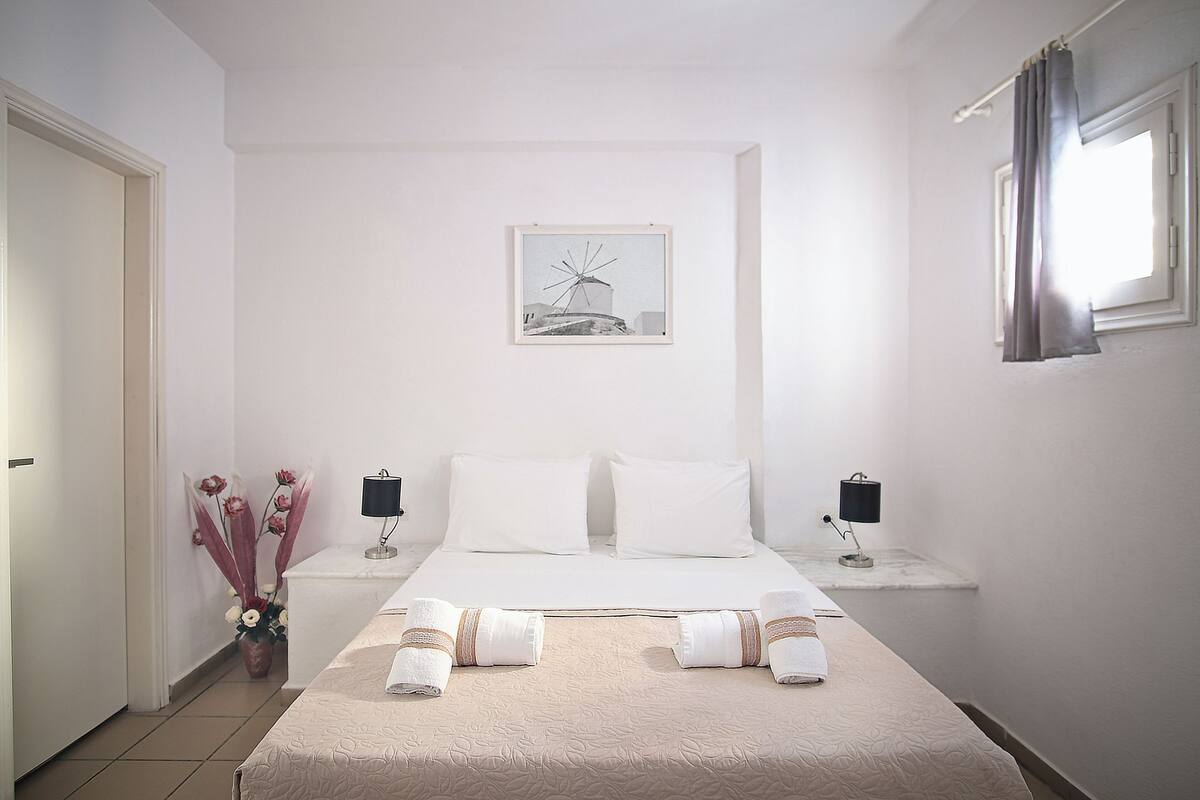 Hotelkamer met een bed, twee nachtkastjes, een raam met gordijnen en een vaas met bloemen. Aan de muur hangt een ingelijst schilderij.
