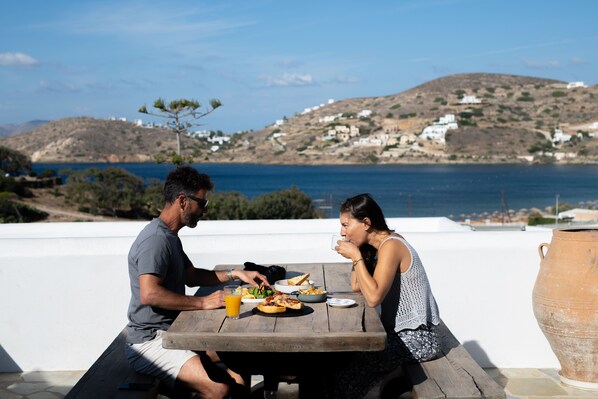 Una pareja cenando en una mesa de madera con vista al mar y colinas al fondo.