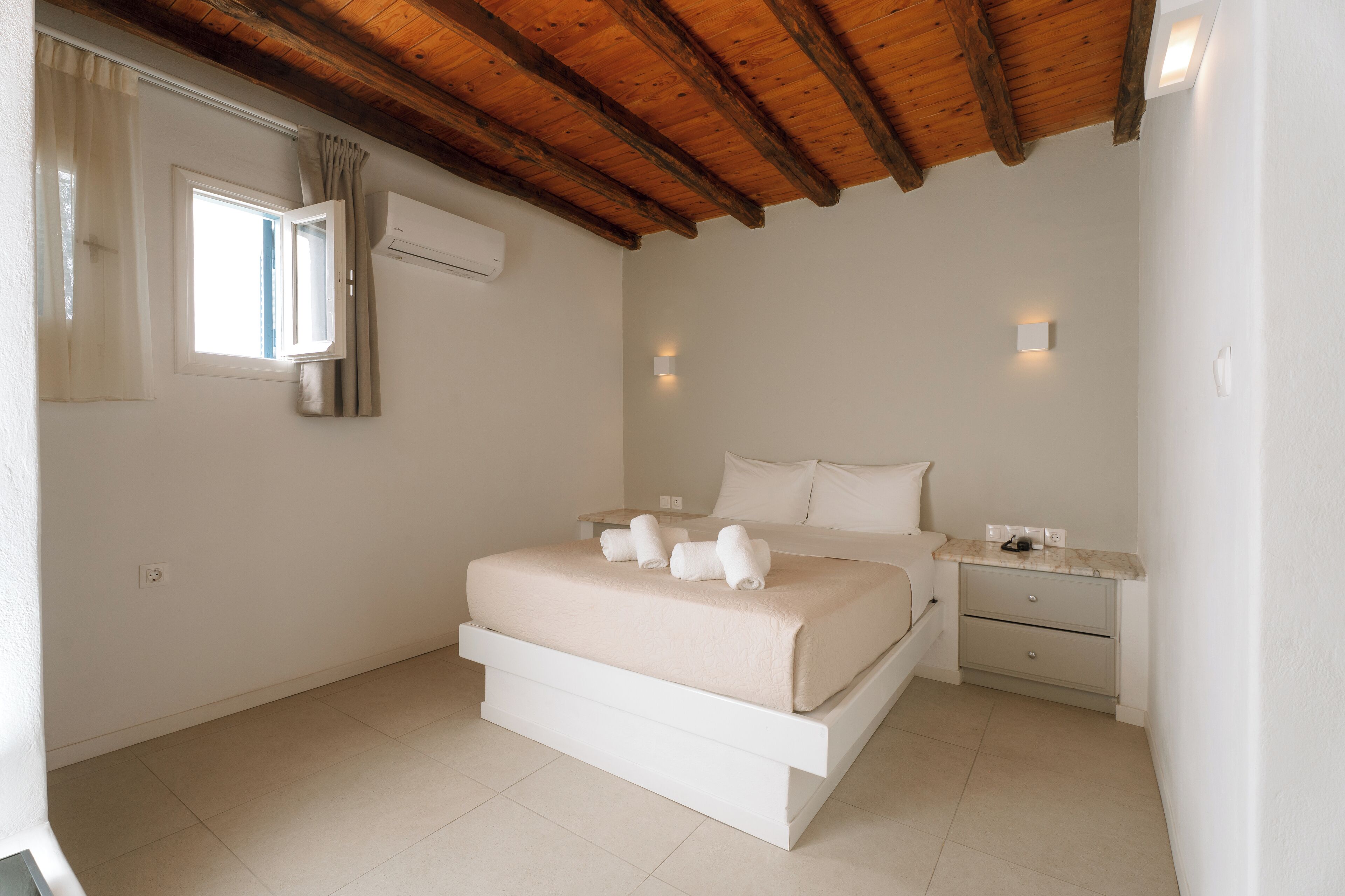 Un dormitorio con techo de madera, cama con ropa de cama blanca, mesita de noche y ventana con cortinas.