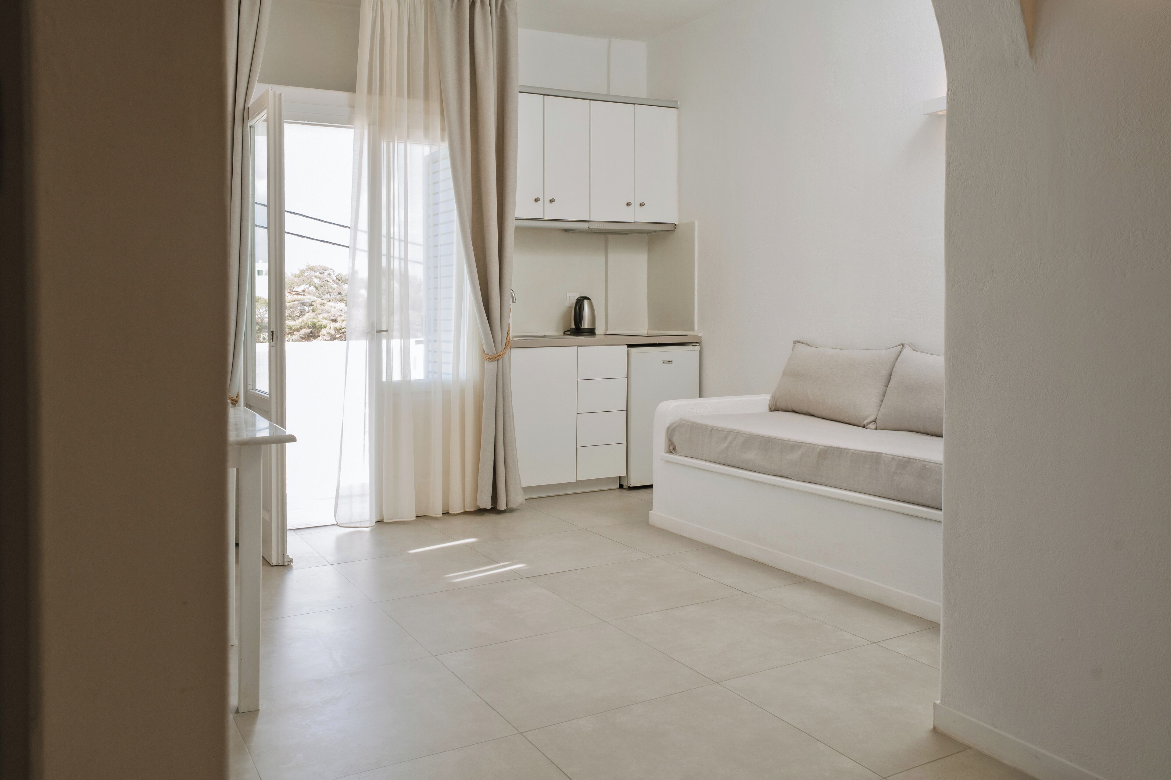 A compact, modern room with a kitchenette, a sofa, and a large window with curtains.