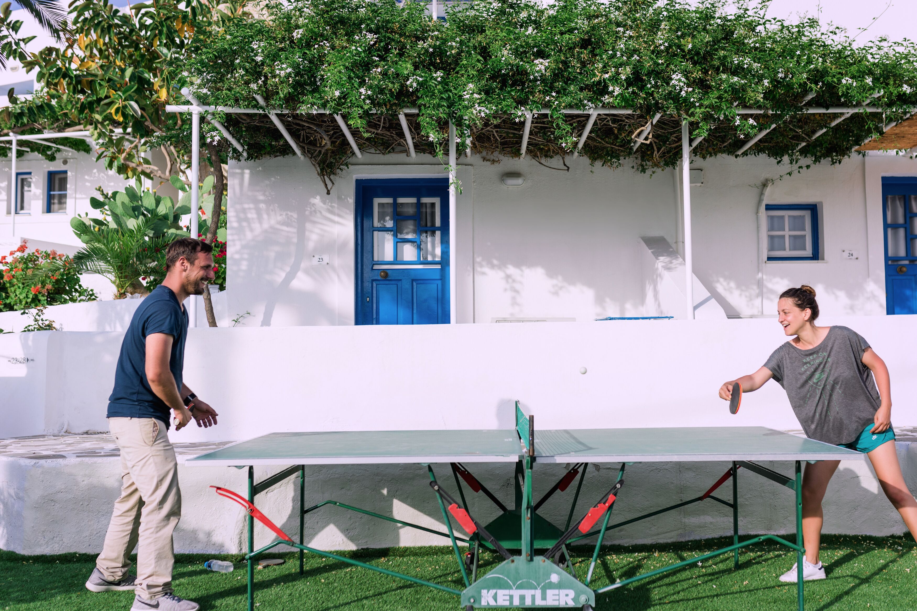 Dos personas jugando ping pong en una cancha al aire libre, con una pared blanca y puertas azules al fondo.