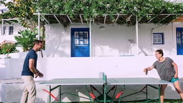 Dos personas jugando ping pong en una cancha al aire libre, con una pared blanca y puertas azules al fondo.