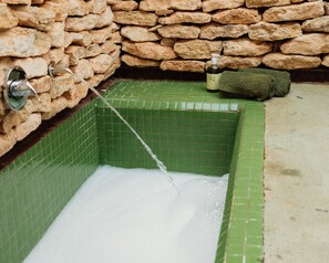 Een bad met groene tegels, een stenen muur, een waterkraan en een fles shampoo.