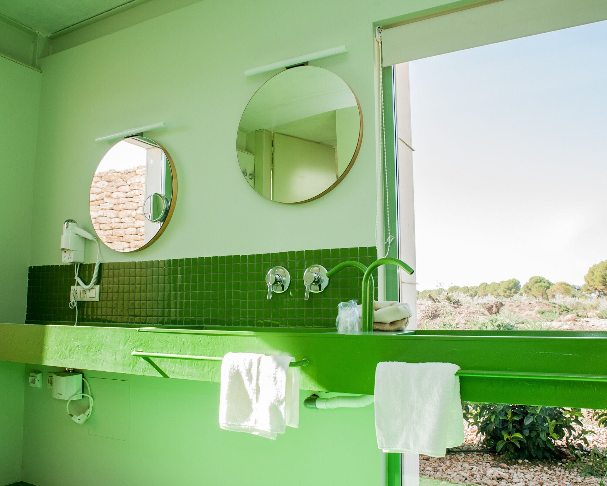 Un baño con dos espejos redondos, un lavabo verde con un toallero y vistas a un paisaje natural a través de una ventana.