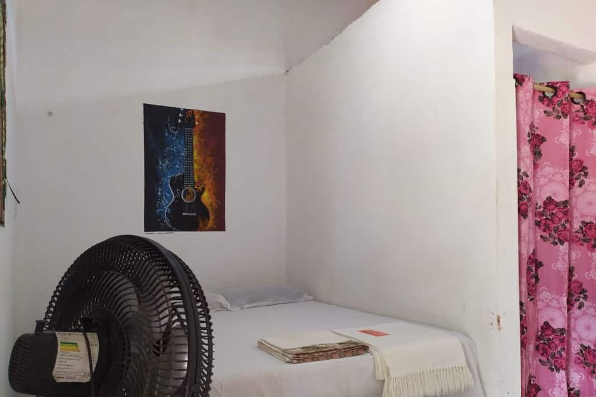 Una habitación pequeña con cama individual, un ventilador, un cartel en la pared y una cortina floral rosa.