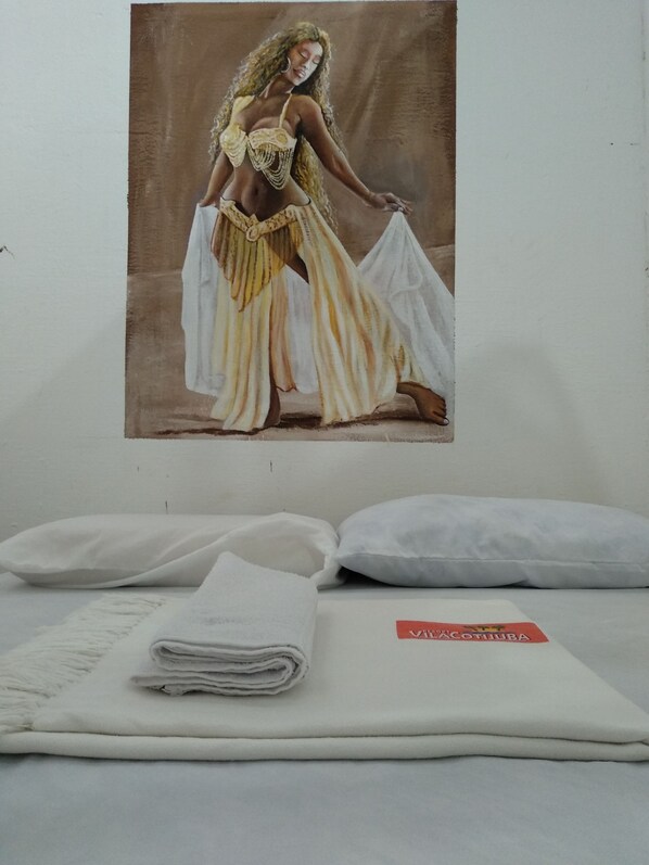 A bed with white linens and pillows, a folded blanket, and a wall-mounted painting of a woman in a yellow and white dress. - Resort Vila Cotijuba (Belém)