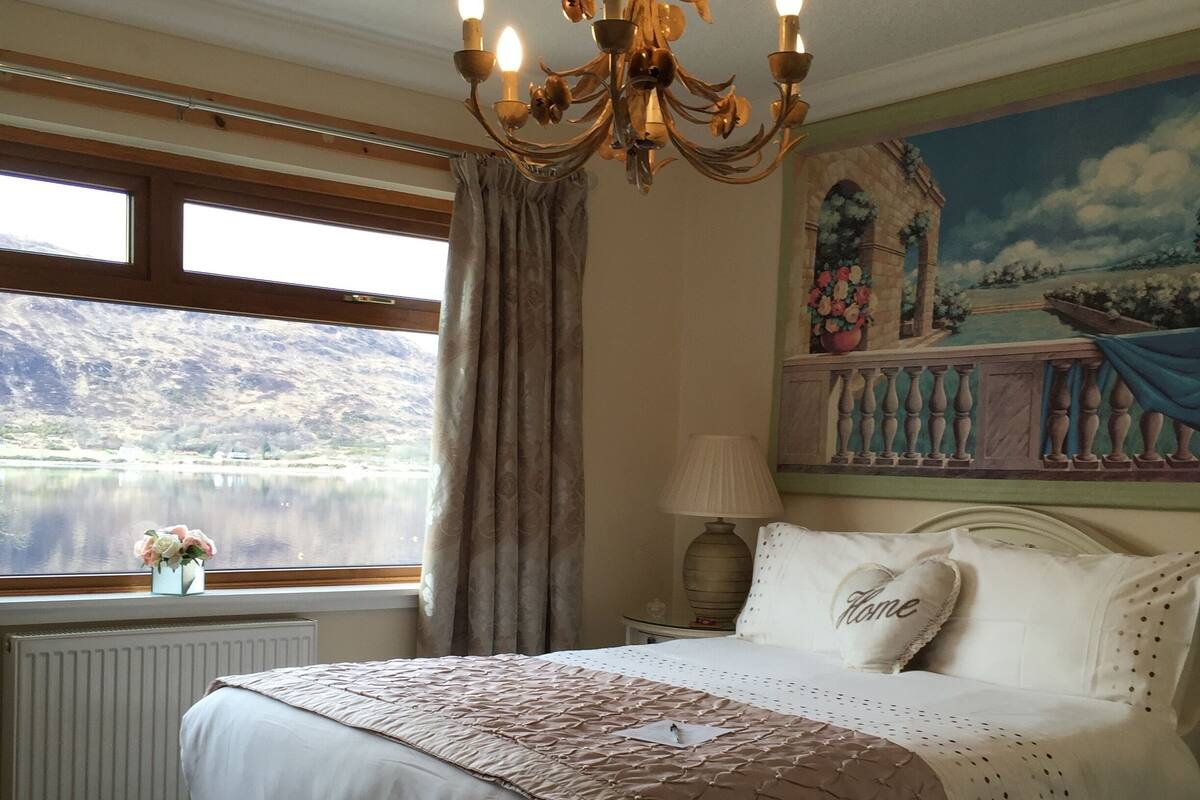 Een slaapkamer met een groot bed, een kroonluchter, uitzicht op een meer en een schilderij van een balkon.