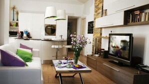 A modern living room with a white sofa, a coffee table with a vase of flowers, a kitchen area with white cabinets, and a television on a wooden stand. - B&B SheLisa (Rome)