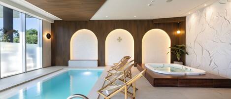 A modern indoor swimming pool with a wooden deck, a hot tub, and a view of the outdoors.