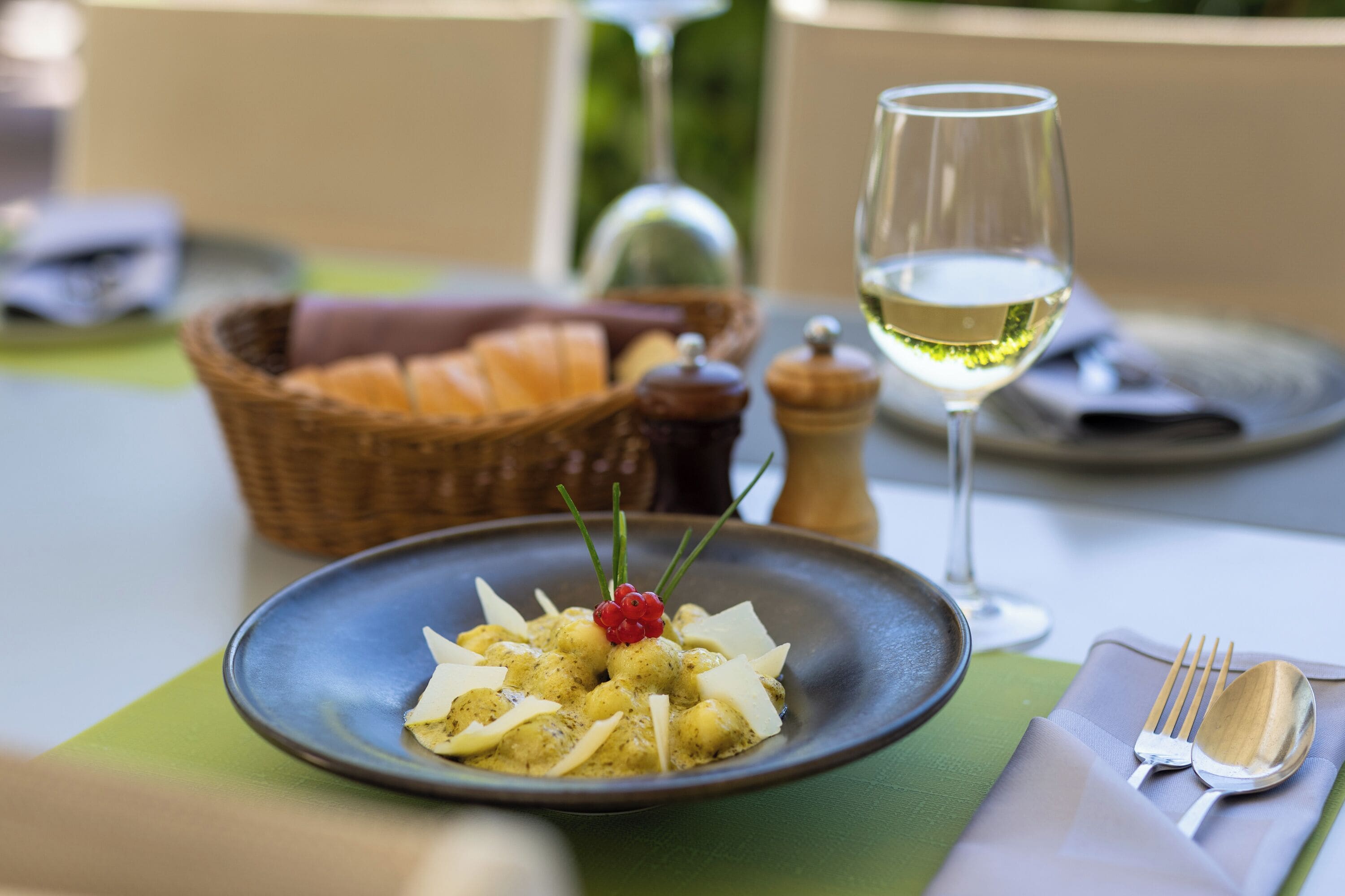 Una mesa de comedor con un plato de comida, una copa de vino blanco y una cesta de pan.
