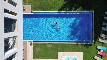 Une vue aérienne d’une piscine où nagent deux personnes, entourée d’une pelouse et de chaises longues.