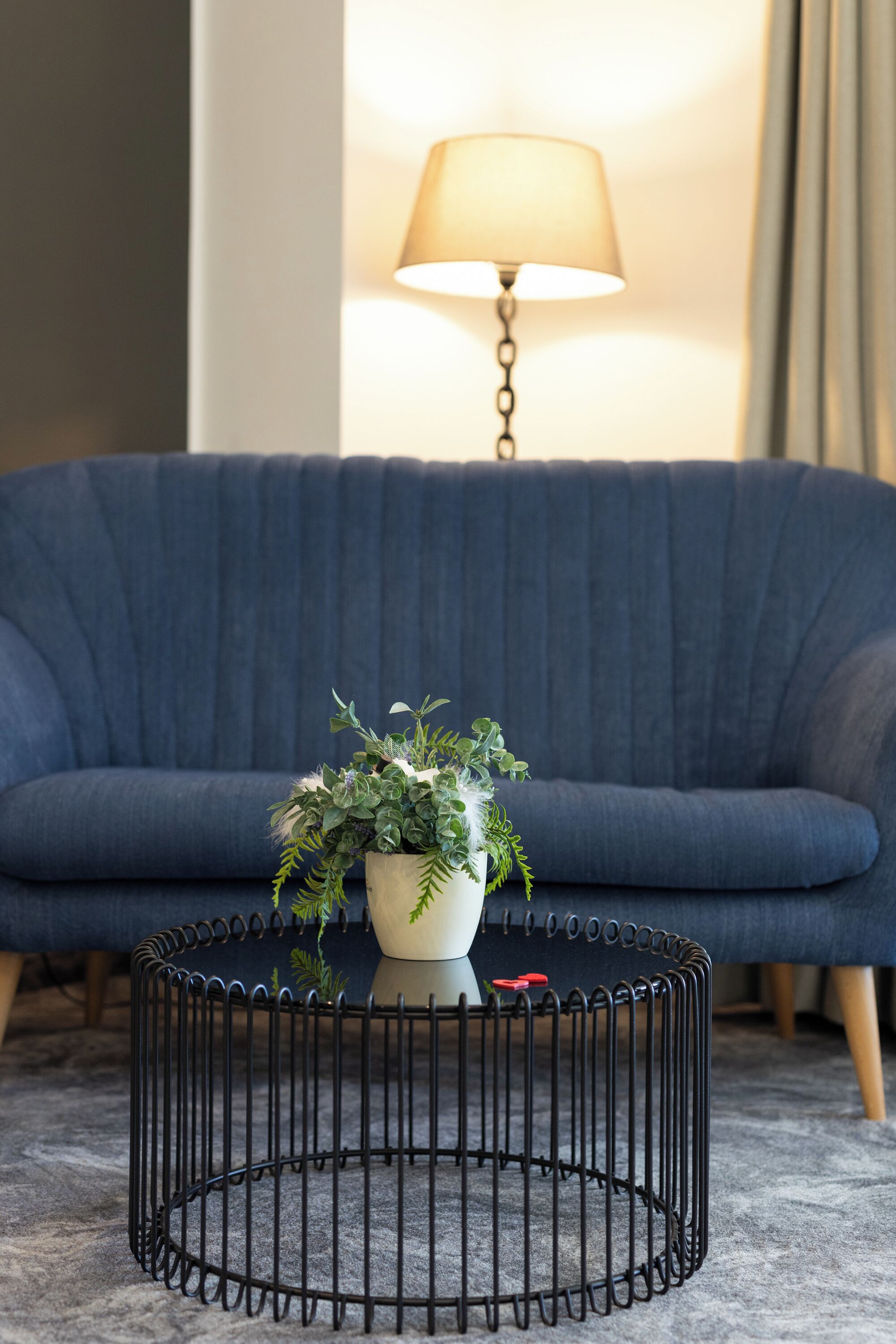 A living room with a dark blue sofa, a round black metal coffee table with a potted plant, and a lamp in the background.