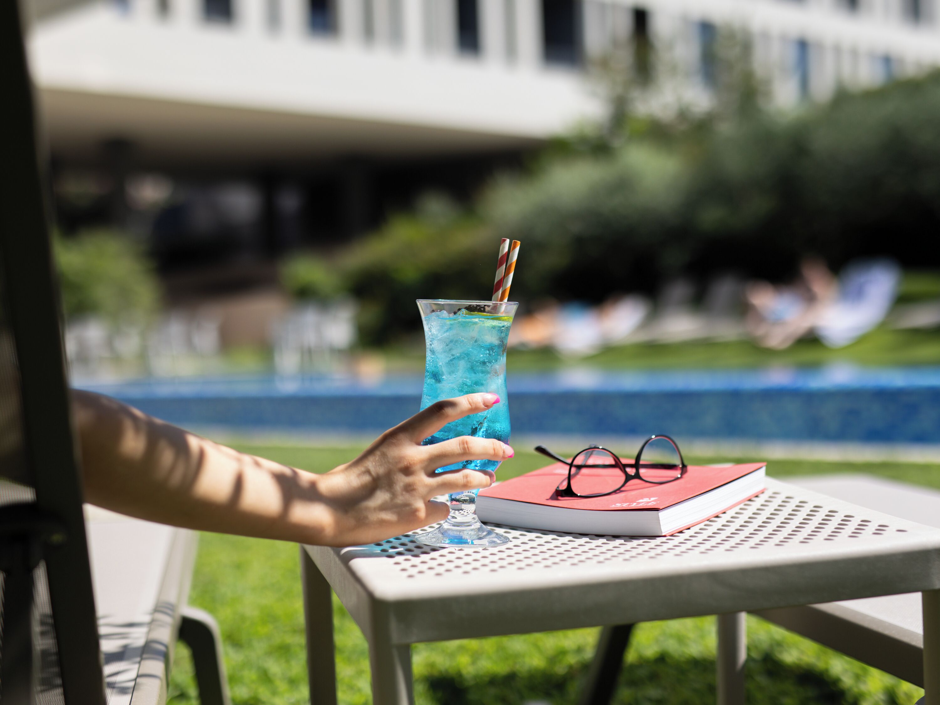 A person holding a glass of blue drink with a striped straw, a book, and a pair of glasses on a table by a pool.