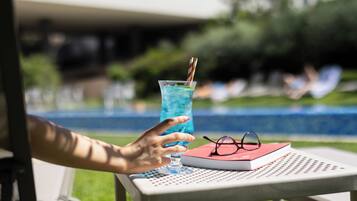 A person holding a glass of blue drink with a striped straw, a book, and a pair of glasses on a table by a pool.