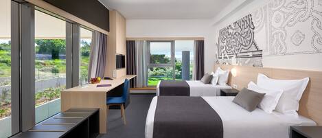 A modern hotel room with two beds, a desk, and a large window offering a view of greenery.