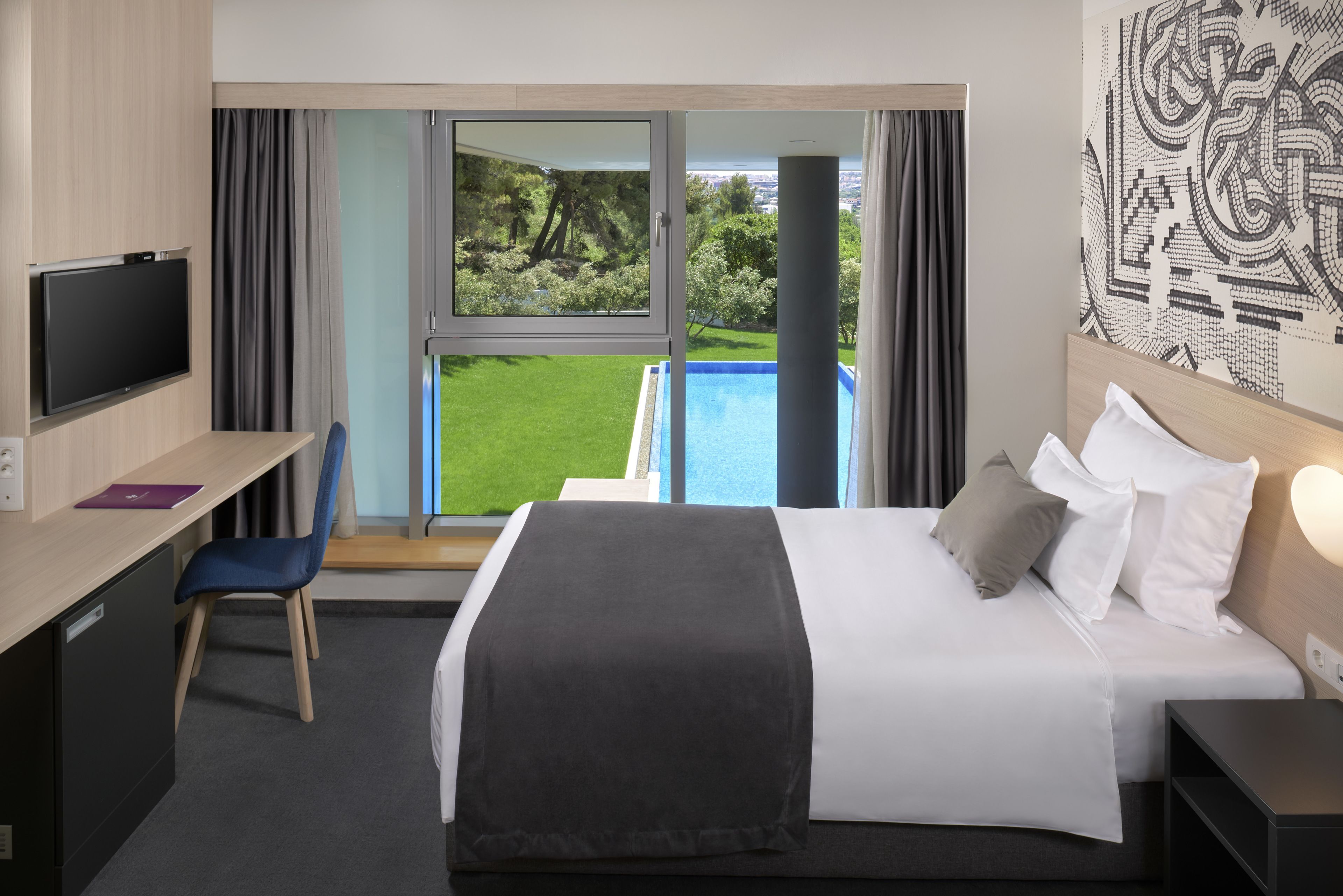 A hotel room with a bed, a desk with a chair, a television, and a large window offering a view of a pool and greenery.