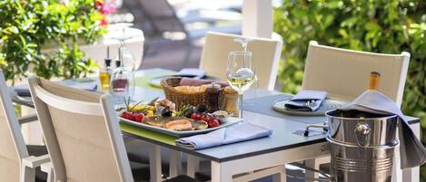 Un espacio para comer al aire libre con sillas blancas y una mesa lista para una comida, una parrilla, y vistas a una piscina y follaje.