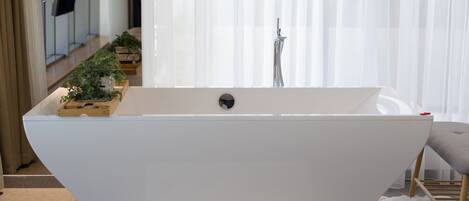 A modern bathroom with a freestanding bathtub, a wooden side table with plants, and a fluffy white rug.