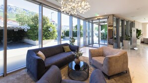 A modern lobby with large windows, a central seating area with sofas and a coffee table, and a decorative chandelier. - Hotel Salona Palace (Solin)