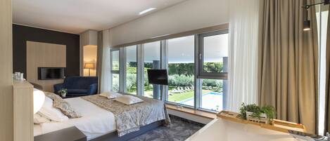 A modern hotel room with a large bed, a sitting area with a chair, a TV mounted on the wall, a large window with a view of greenery, and a bathtub.
