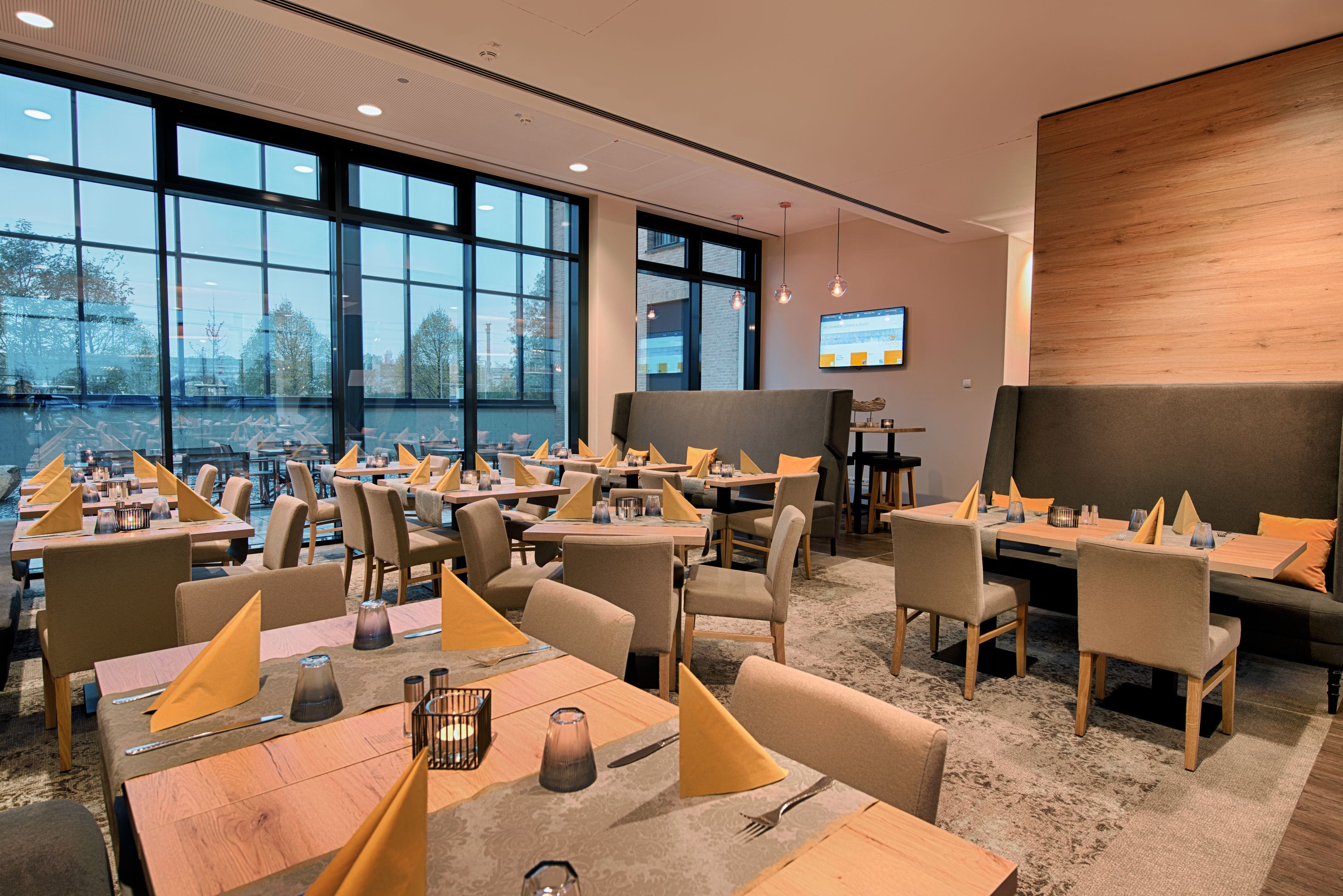 A modern dining area with multiple tables and chairs, large windows, and a flat-screen TV on the wall.
