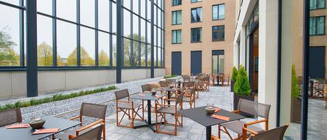 A spacious outdoor seating area with multiple tables and chairs, large windows, and a view of a building with multiple floors.