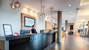 A modern reception area with a receptionist, a large screen displaying information, and a counter with gaming consoles.