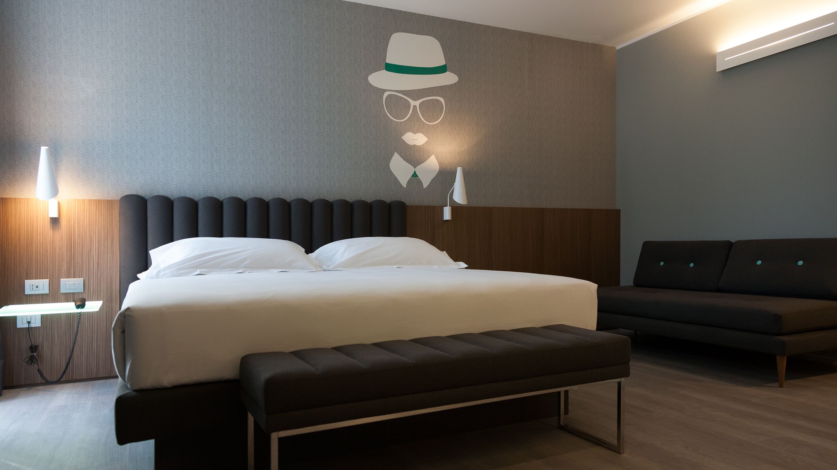 A modern hotel room with a large bed, a bench, a sofa, and a wall-mounted light fixture.