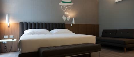 A modern hotel room with a large bed, a bench, a sofa, and a wall-mounted light fixture.