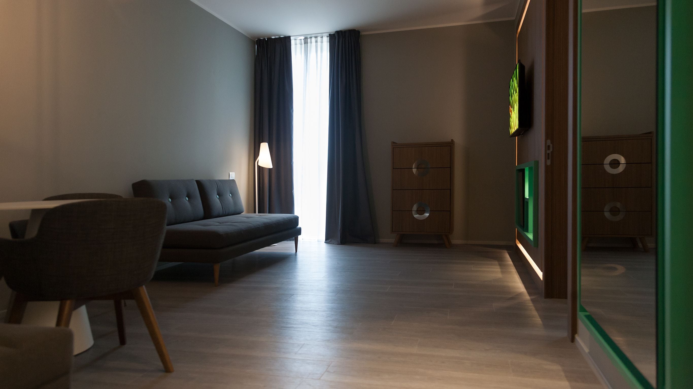 A modern, well-lit room with a sofa, a chair, a desk, and a TV mounted on the wall.