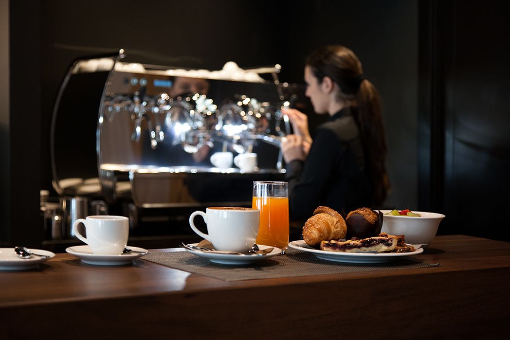 A breakfast setup with a coffee machine, two cups of coffee, a glass of orange juice, croissants, and a bowl of fruit salad.