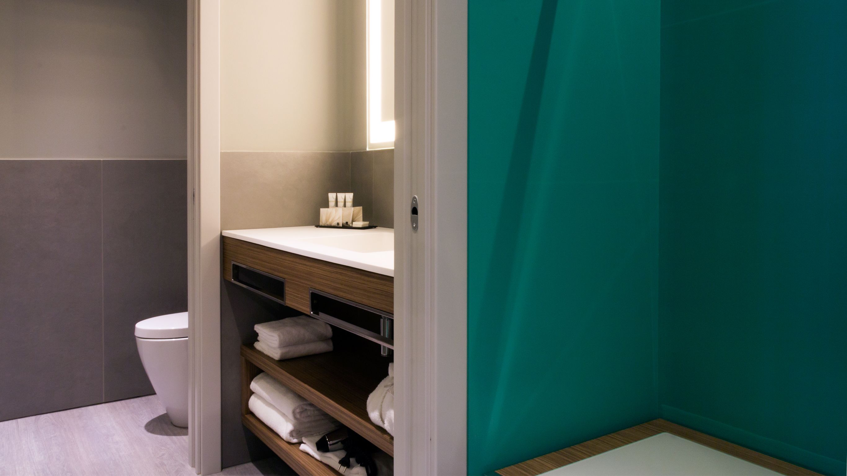 A modern bathroom with a white toilet, a sink with a mirror, and a shelf with towels.