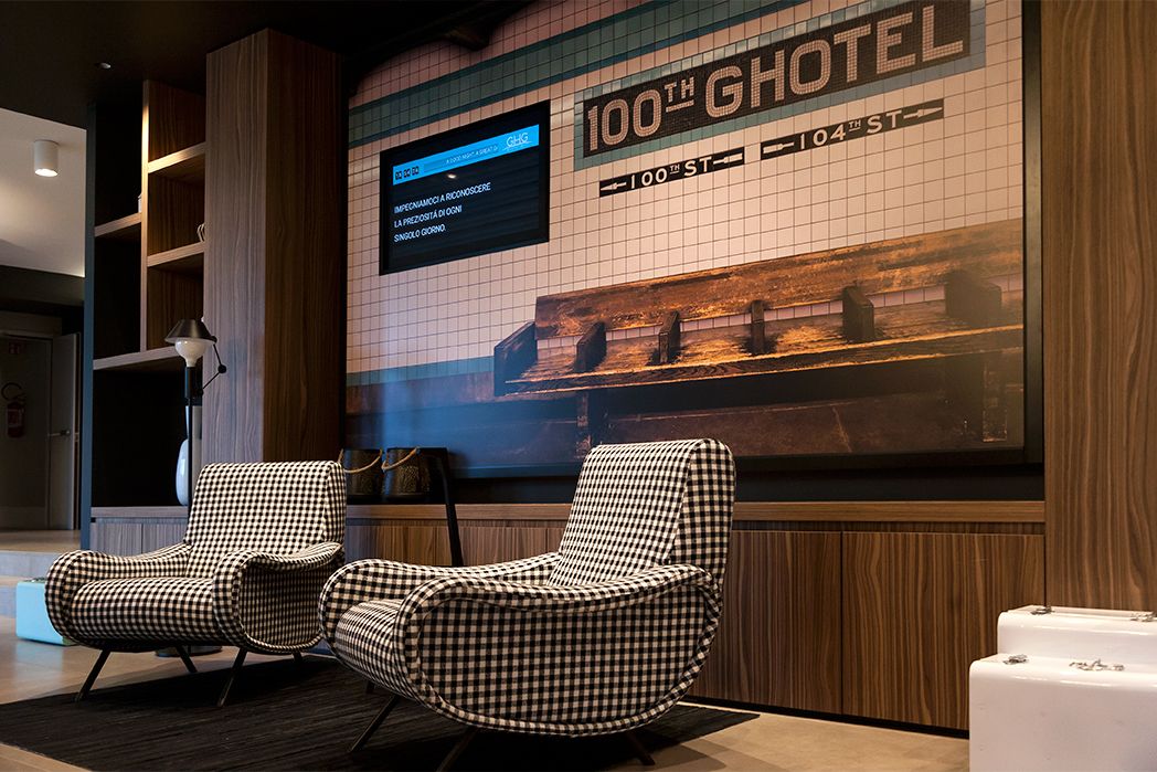 A lobby with checkered armchairs, a digital display with hotel information, and a tiled wall with the hotel's name.