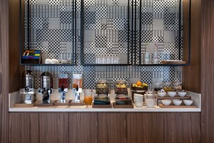 Una barra de desayuno con diversas opciones de comida y bebida, incluyendo un bol de fruta, cereales y una máquina de café.