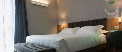 A neatly made bed with white linens, a grey bench, and a wall-mounted lamp.