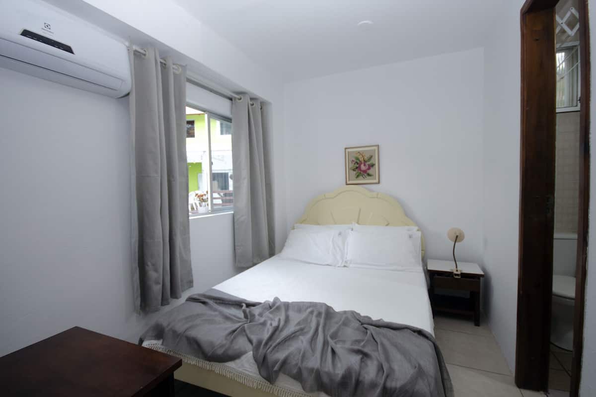 A bedroom with a bed, a bedside table, a window with curtains, and a wall-mounted air conditioner.