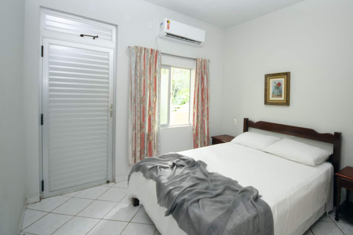 A bedroom with a bed, a window with curtains, an air conditioning unit, a bedside table, and a picture on the wall.