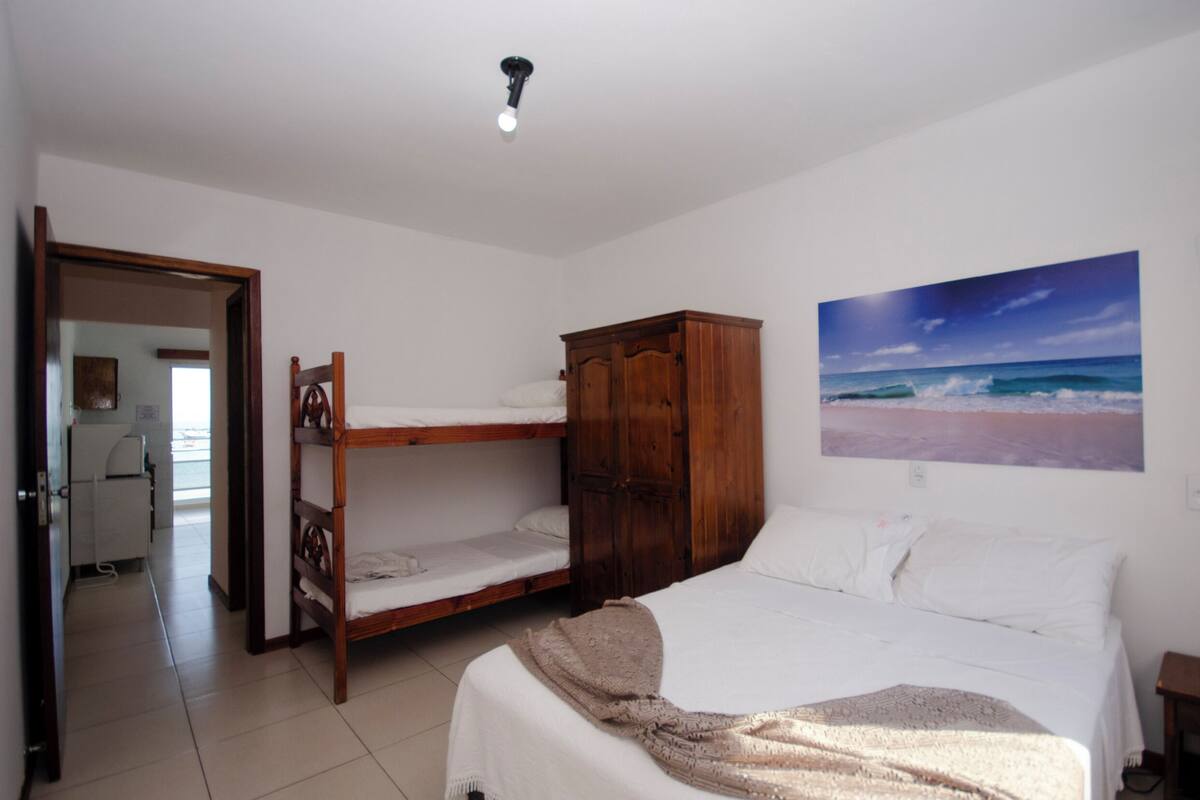 A bedroom with bunk beds, a wooden wardrobe, a bed with a blanket, and a beach painting on the wall.
