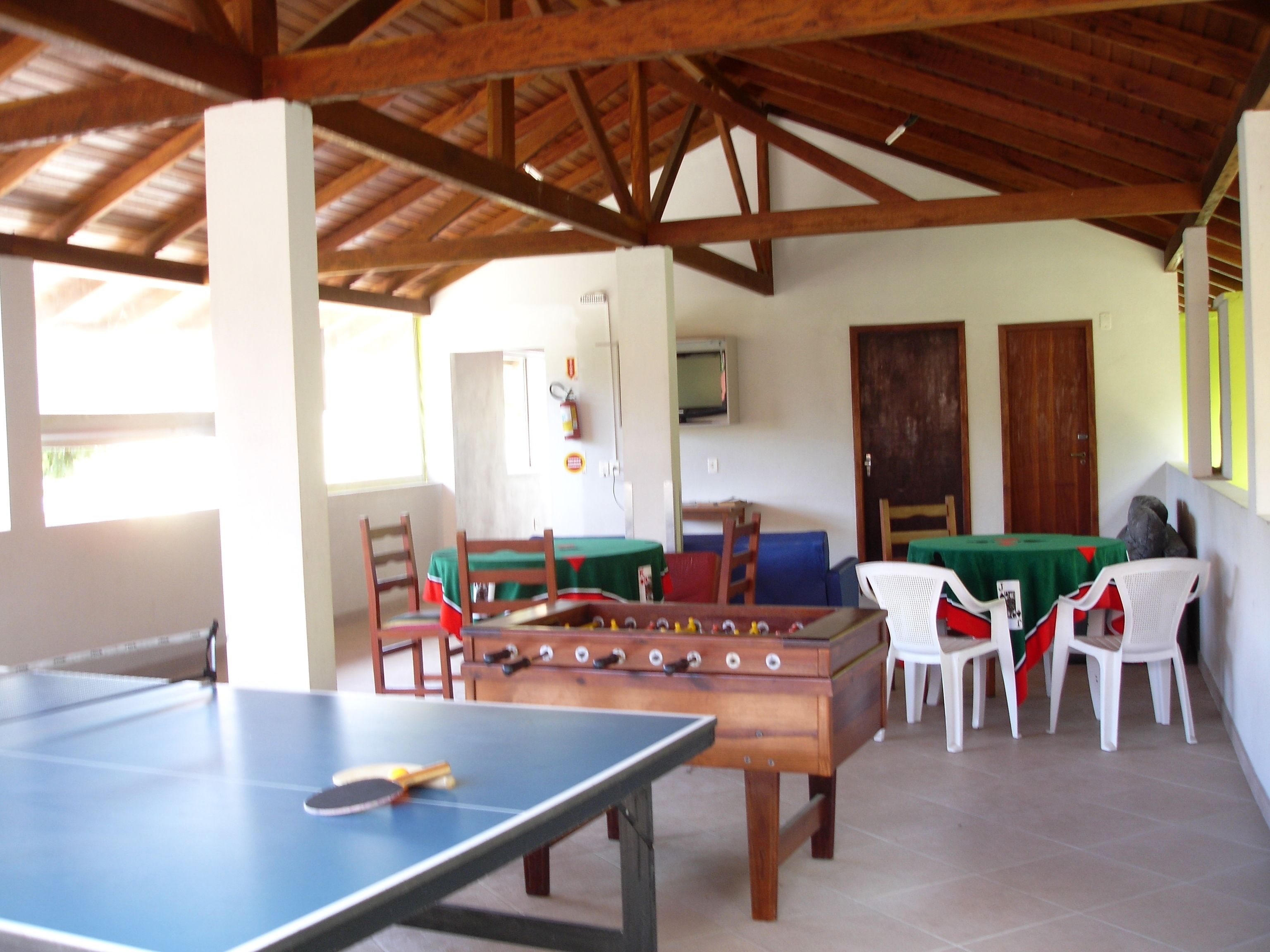 A room with a ping pong table, a foosball table, and several chairs. There is a television on the wall and a door in the background.