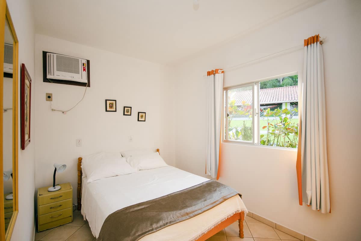 A bedroom with a bed, a bedside table with a lamp, a window with curtains, and an air conditioning unit on the wall.