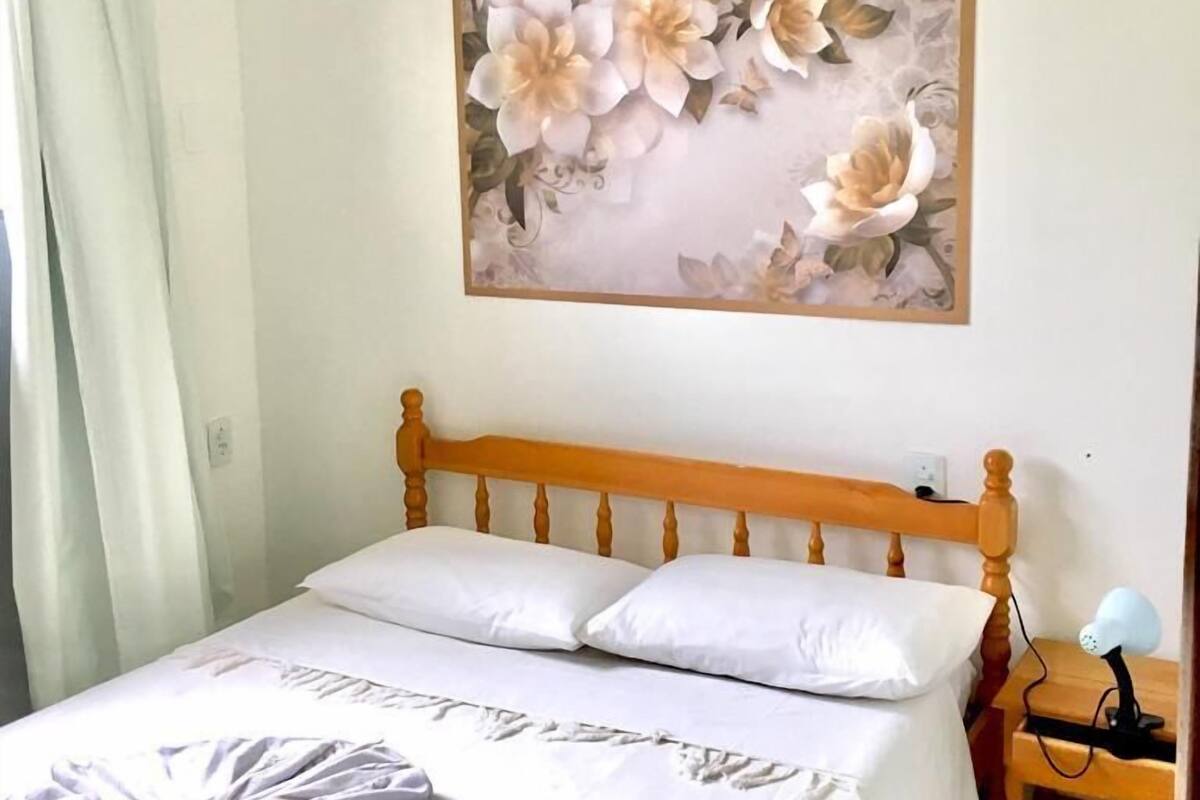 A bedroom with a wooden bed, white bedding, a wall-mounted air conditioner, a floral artwork, and a bedside lamp.