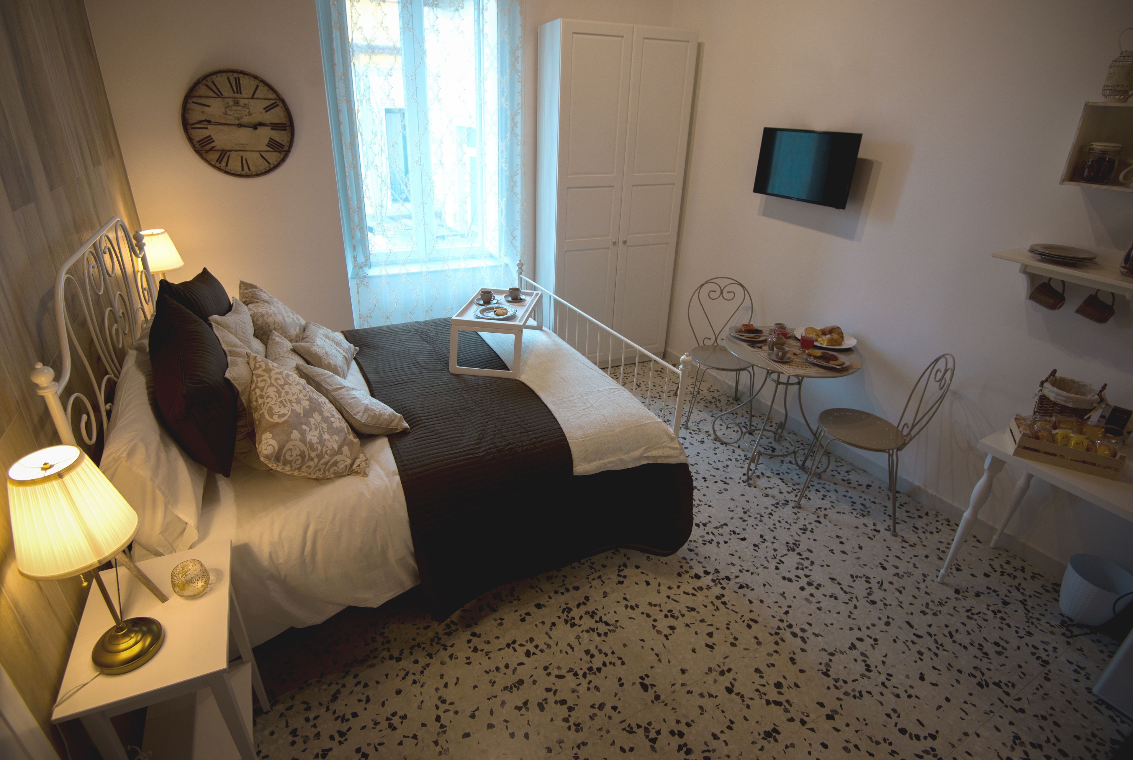 Une chambre à coucher comprenant un lit, une table de chevet, une lampe, une horloge, un téléviseur et un petit coin repas avec une table et des chaises.