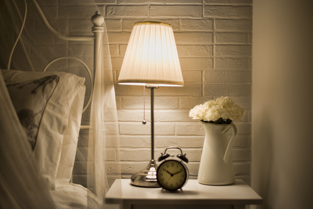 Ein Nachttisch mit Lampe, Wecker und Blumen in einer Vase.