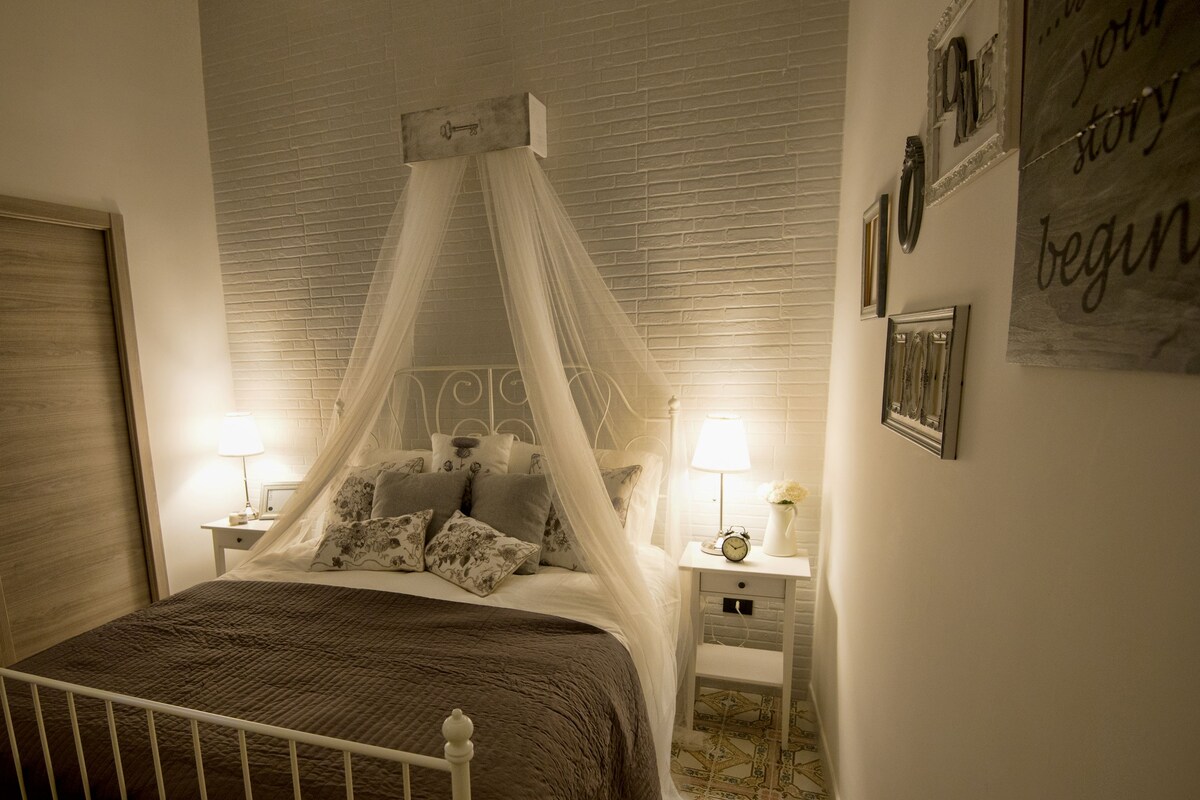 A bedroom with a four-poster bed, bedside tables with lamps, a wooden door, and a wall with framed pictures and a sign.