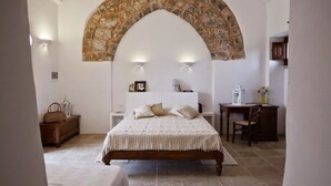 A bedroom with a stone archway, a bed with a white bedspread, a nightstand, a desk, and a chair. - Masseria Cisternella (Alliste)