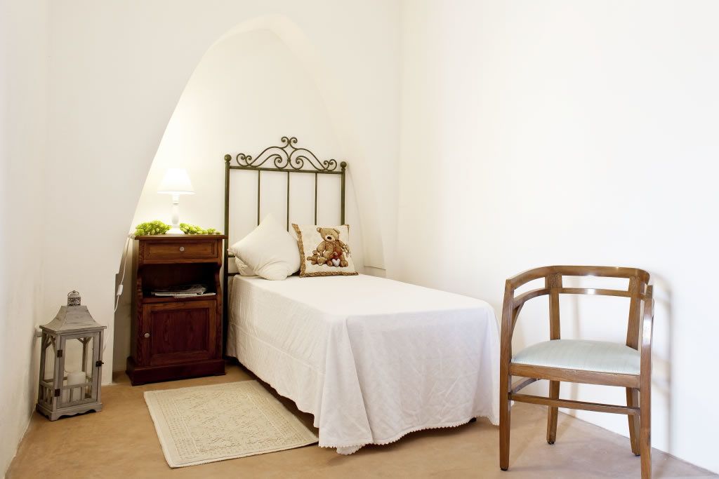 A bedroom with a single bed, a wooden nightstand, a small table with a lamp, a chair, and a decorative item on the bed.