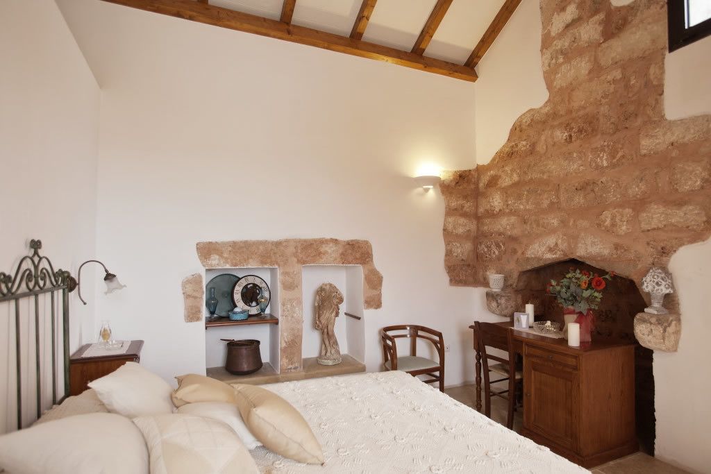 A bedroom with a stone fireplace, a bed with white linens, a wooden desk, and a shelf with decorative items.